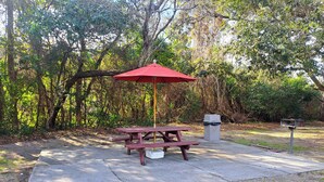 A picnic table with a red umbrella, a concrete ground, and a trash bin in a wooded area.