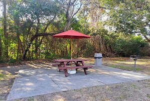 A picnic table with a red umbrella, a concrete ground, and a trash bin in a wooded area.