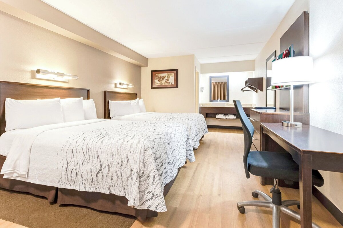 A hotel room with a large bed, a desk with a chair, a mirror, and a wall-mounted lamp.
