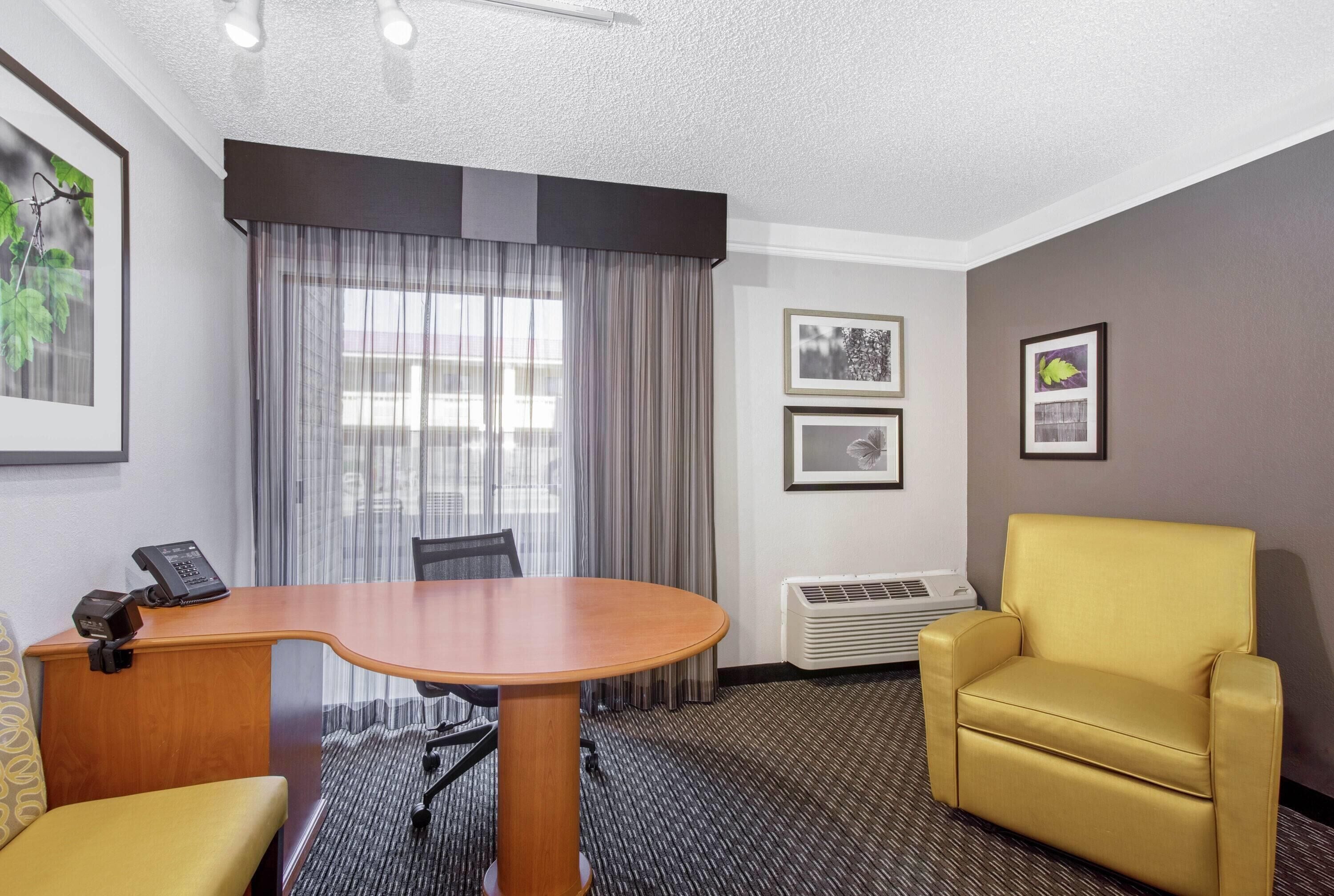 Habitación de hotel con un escritorio de madera, un sillón amarillo, un pequeño sofá, un teléfono y cuadros en la pared.