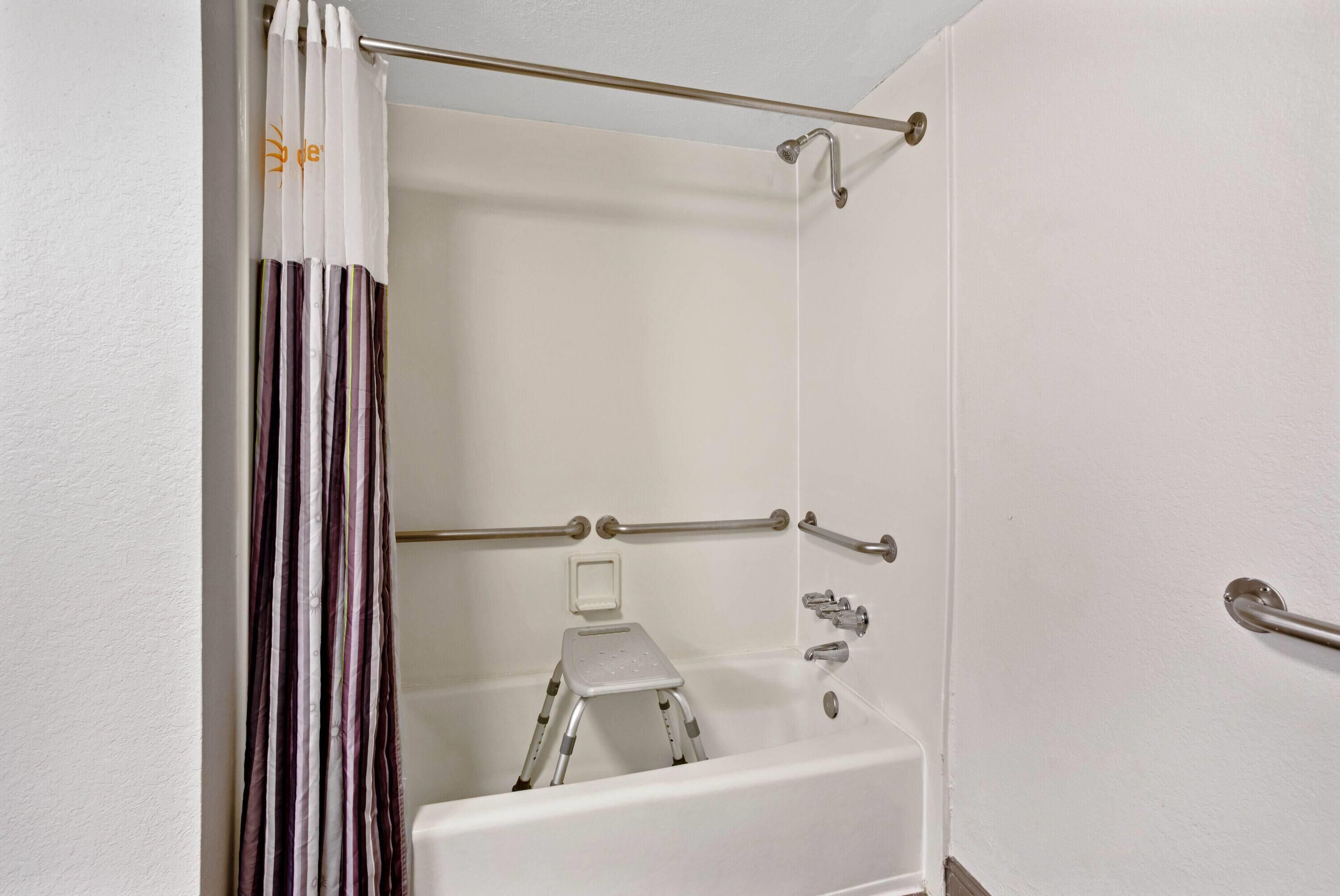 Un baño con una bañera blanca, barras de apoyo, una cortina de ducha y un taburete.