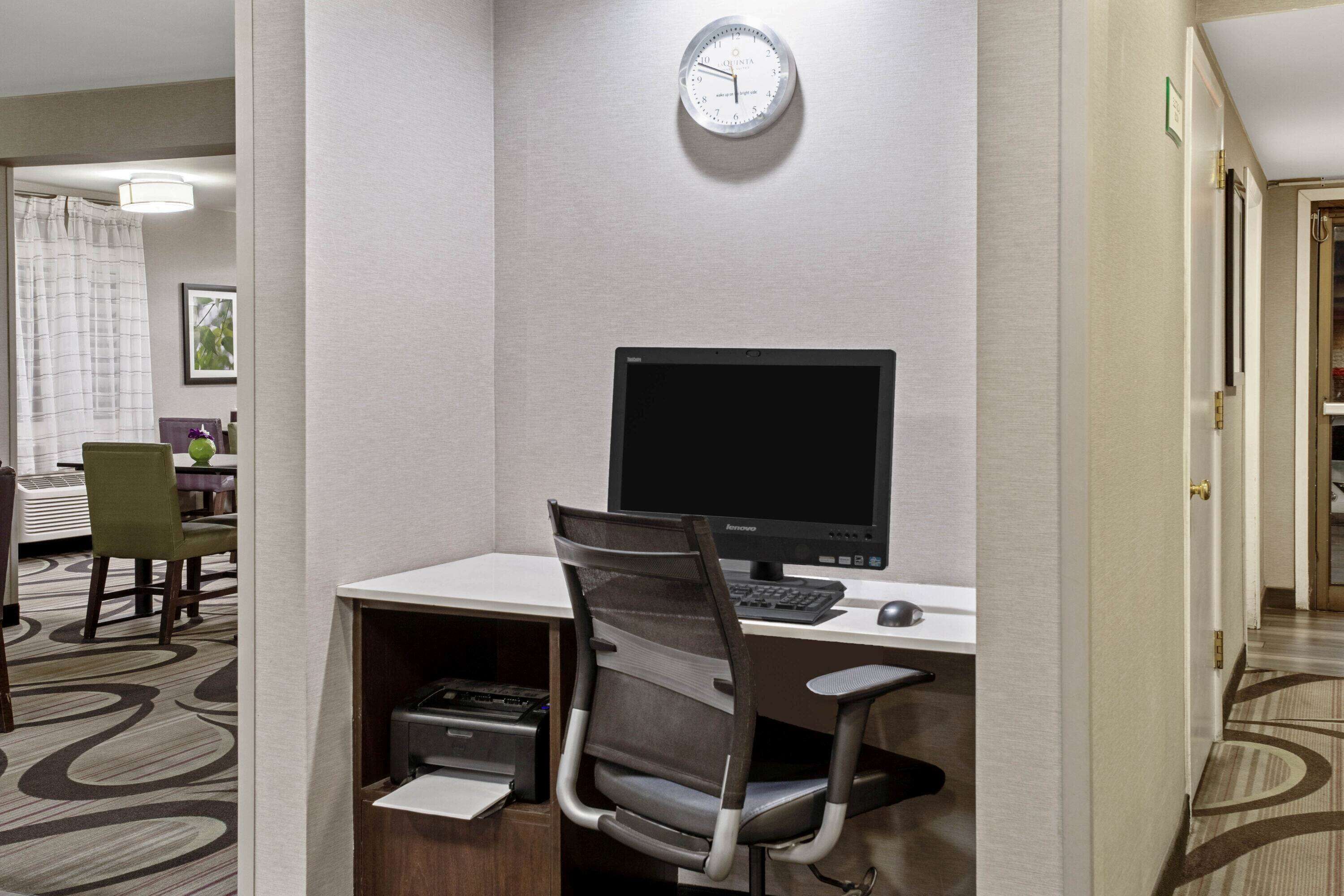 A hotel room with a desk, computer, chair, and a clock on the wall.
