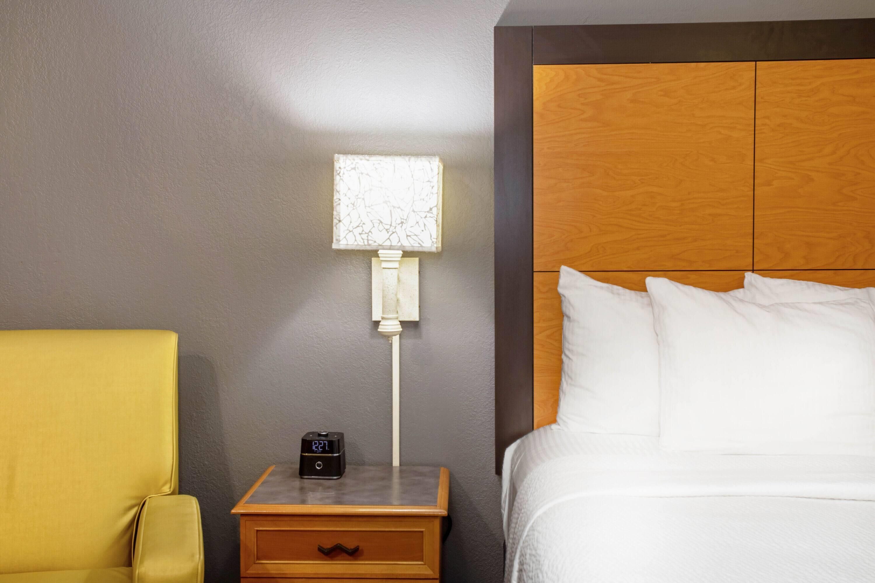 A hotel room with a wooden headboard, a bedside table with a clock, a lamp, and a yellow armchair.