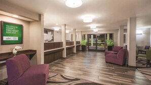 Un vestíbulo de hotel con mostrador de recepción, asientos morados, un letrero verde y una planta.