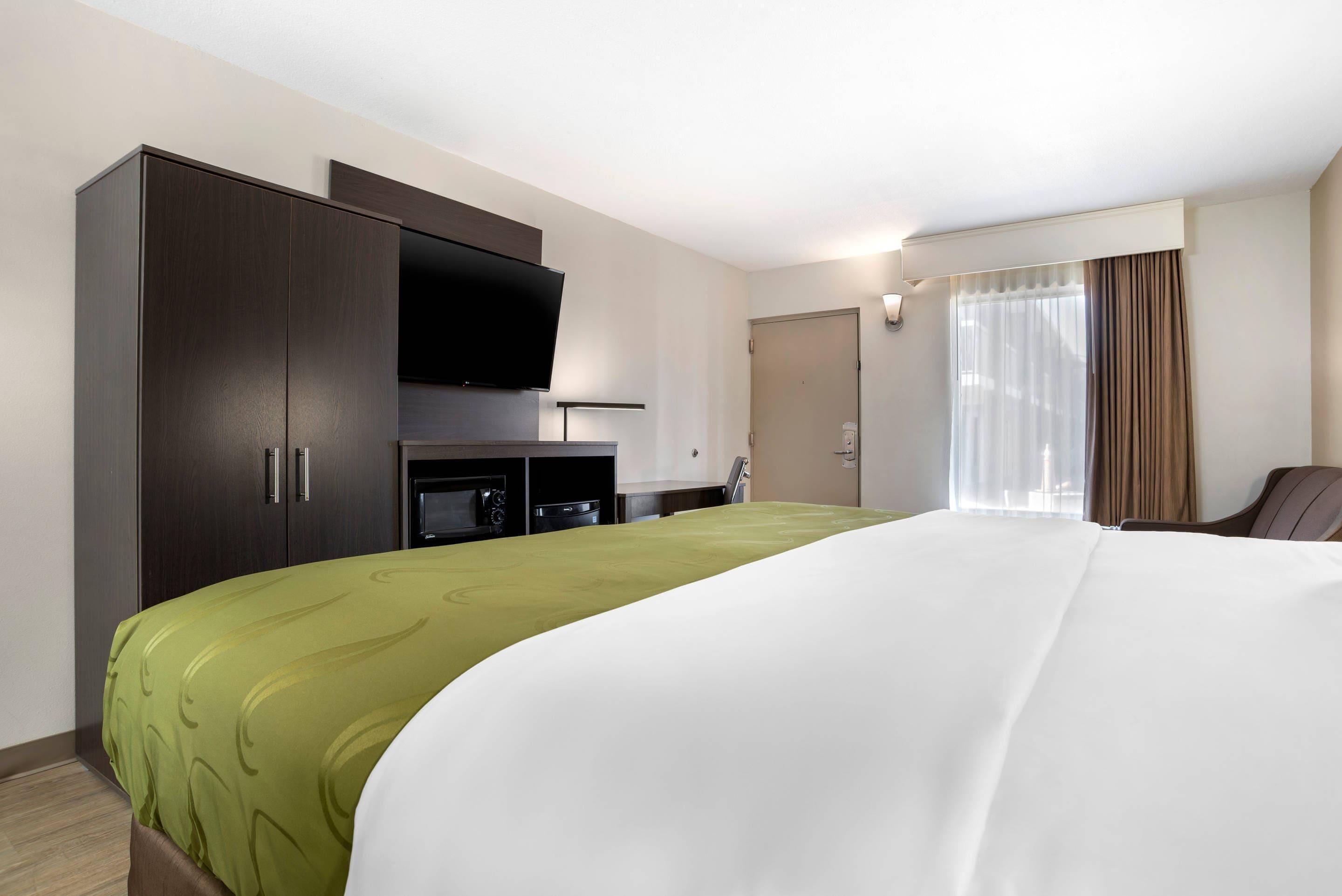 Habitación de hotel con una cama grande, televisor montado en la pared, minibar y una ventana con cortinas.