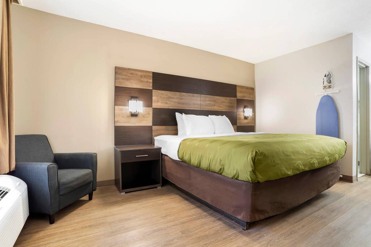 Una camera d'hotel con una testiera in legno, un letto con coperta verde, un comodino, una sedia e un asse da stiro fissato alla parete.