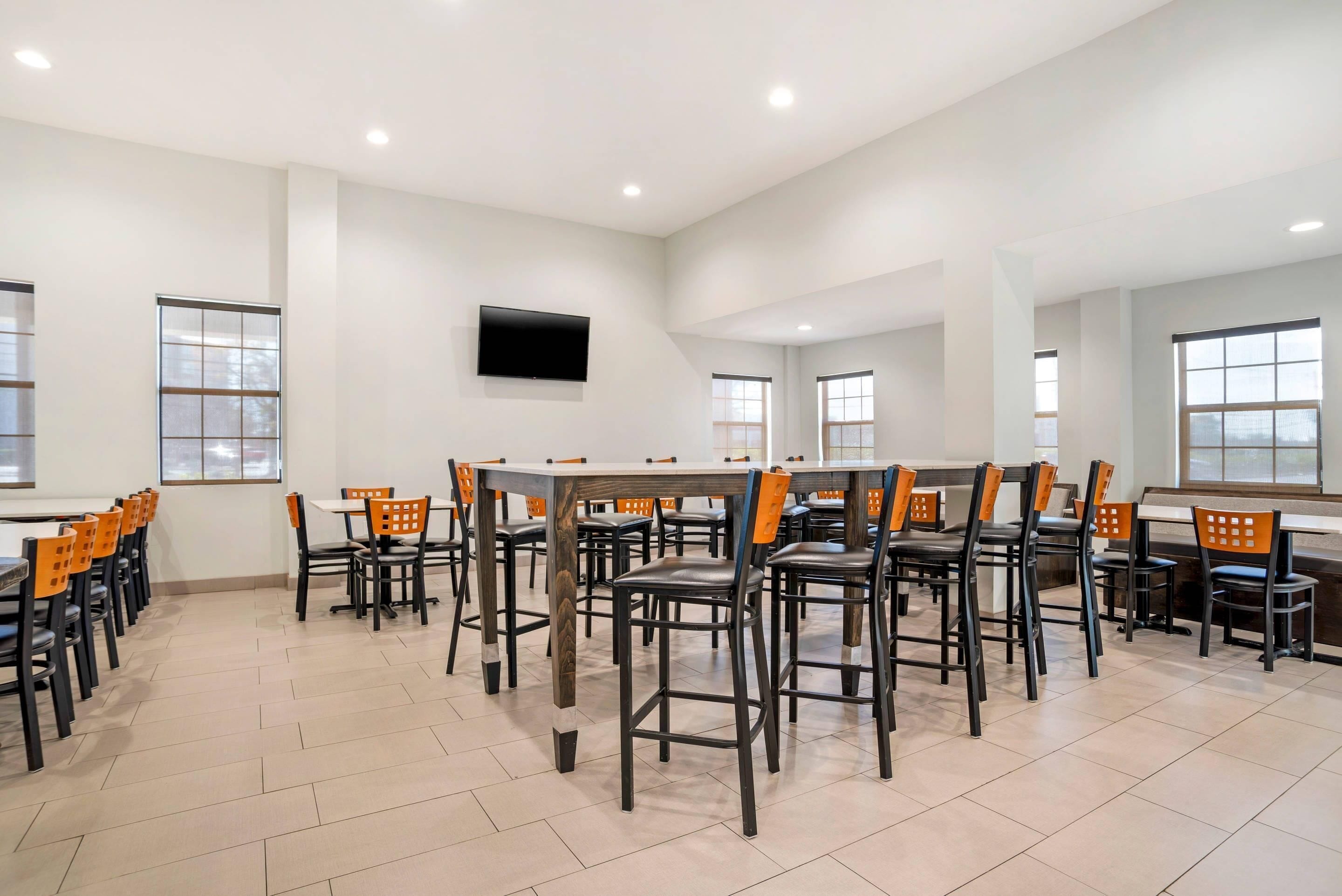 A spacious dining area with multiple tables and chairs, a flat-screen TV mounted on the wall, and large windows providing natural light.
