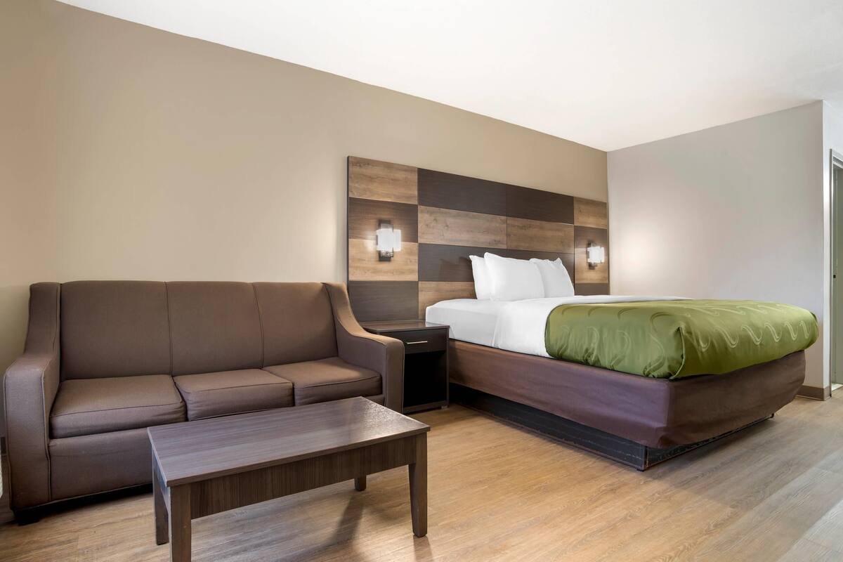 Una camera d'albergo con un divano marrone, un tavolino in legno, un letto con una coperta verde e un punto luce a parete.