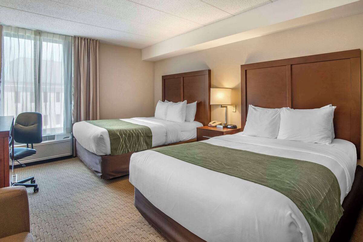 A hotel room with two beds, a desk, a chair, a lamp, and a window with curtains.