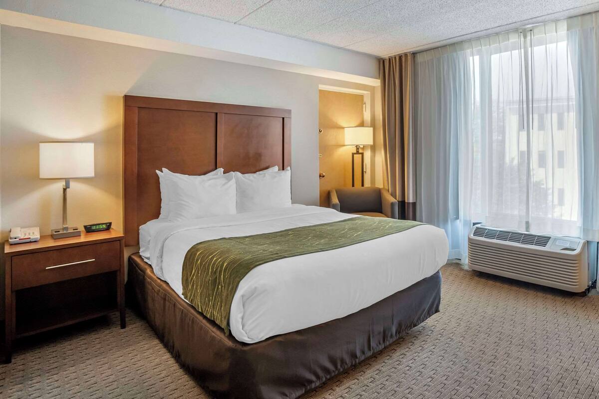 A hotel room with a large bed, a nightstand with a lamp, a chair, and an air conditioning unit.