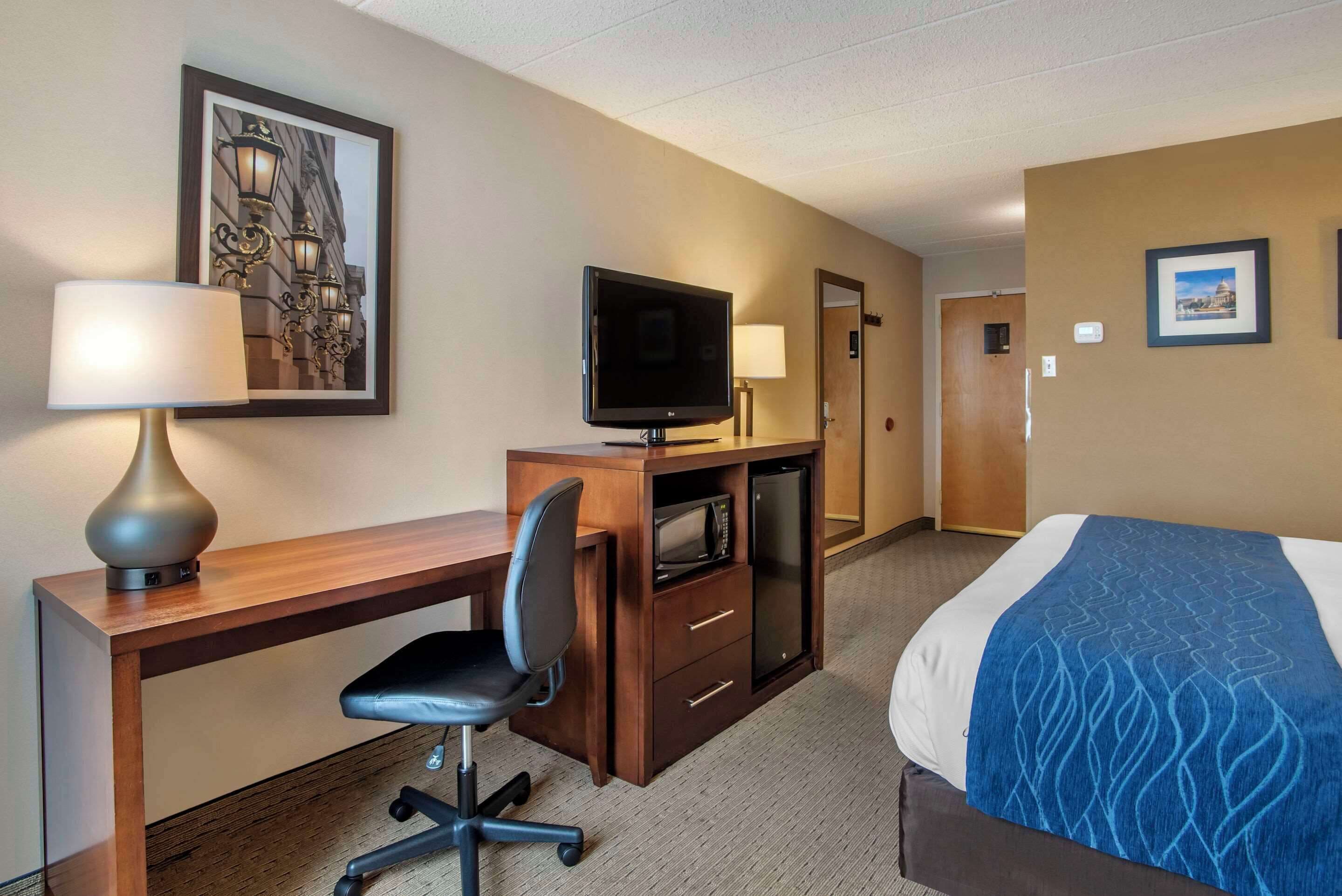 Una habitación de hotel con un escritorio de madera, televisión, una silla, una cama con manta azul y un cuadro en la pared.