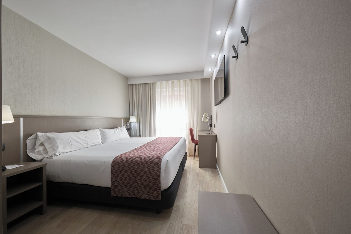 A hotel room with a large bed, bedside tables, a desk, a chair, and a TV mounted on the wall.