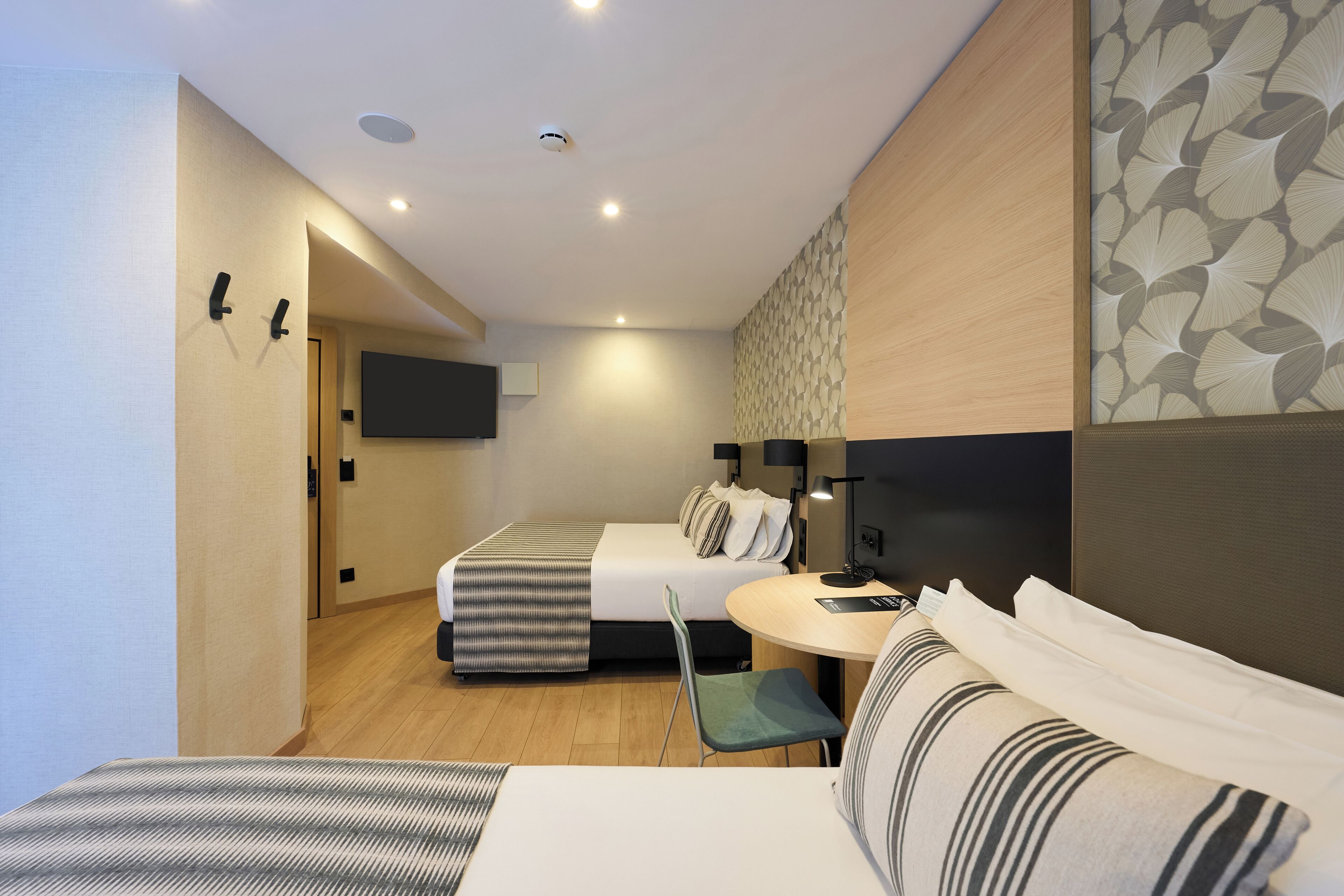 A hotel room with a bed, a desk, a chair, a TV mounted on the wall, and a patterned wall.