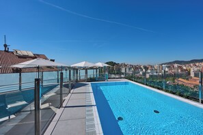 A rooftop pool with clear blue water, surrounded by glass railings and white umbrellas, overlooking a cityscape. - Catalonia Park Güell (Barcelona)