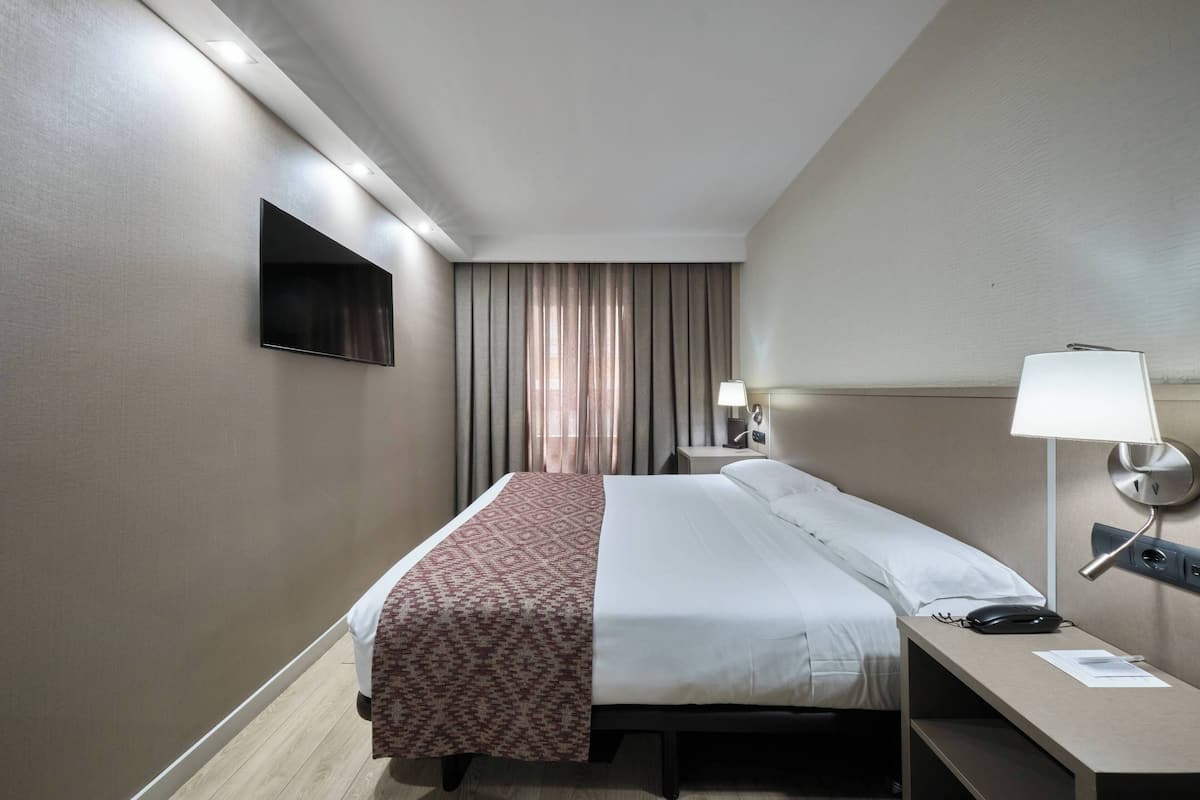 A hotel room with a bed, bedside table, lamp, and a TV mounted on the wall.