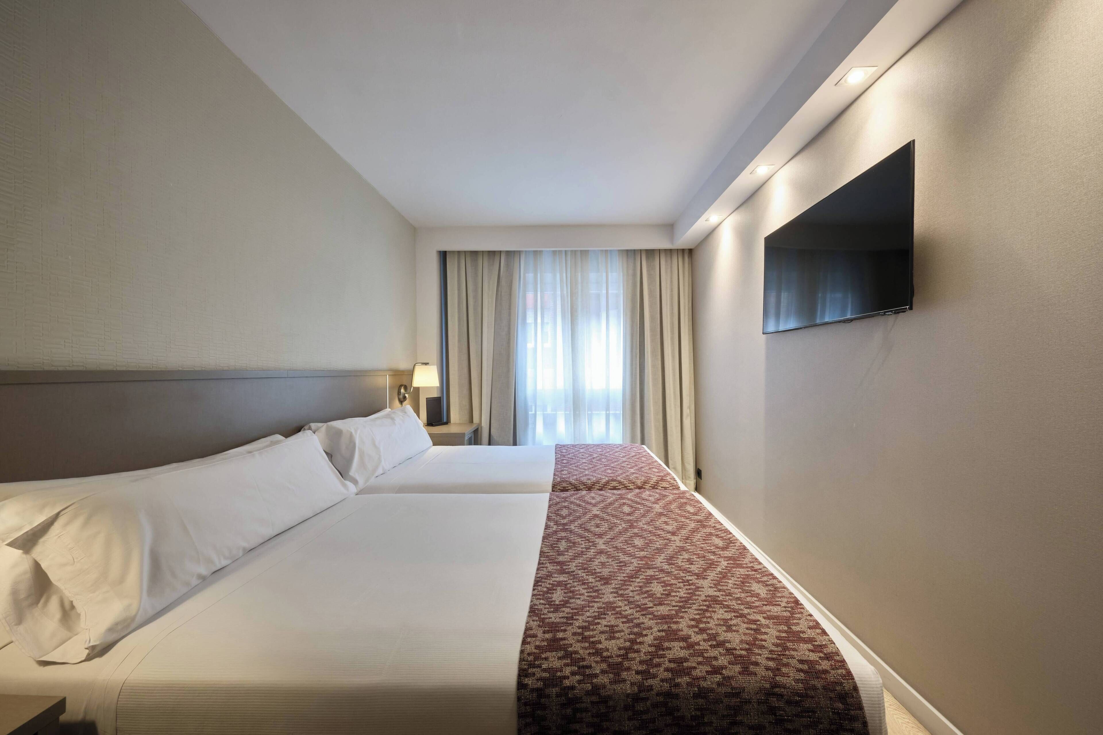Ein Hotelzimmer mit zwei Betten, einem an der Wand montierten Flachbildfernseher und einem Fenster mit Vorhängen.