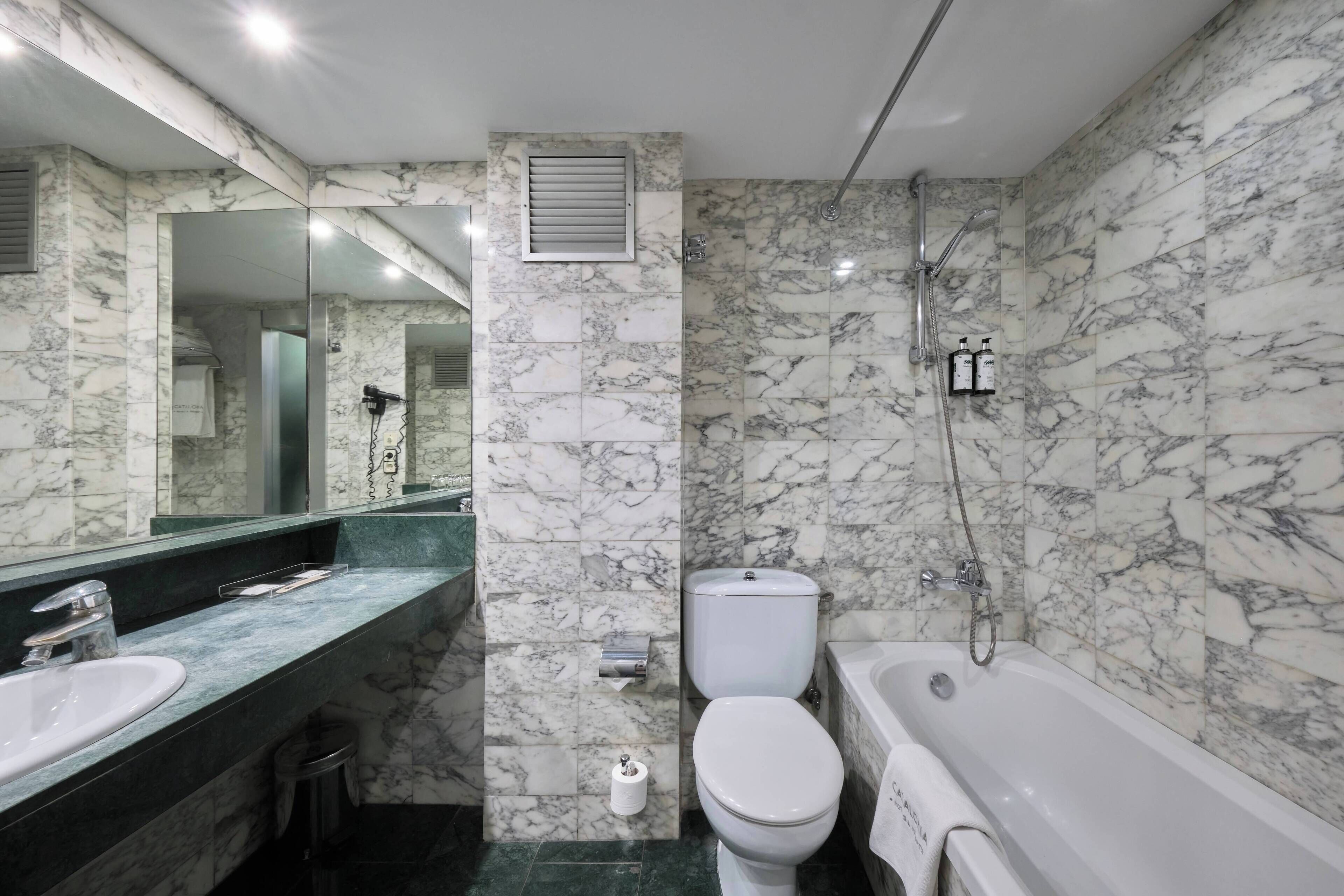 Un baño con lavamanos, inodoro y bañera de mármol, con un espejo grande y cabezal de ducha.