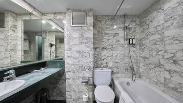Un baño con lavamanos, inodoro y bañera de mármol, con un espejo grande y cabezal de ducha.