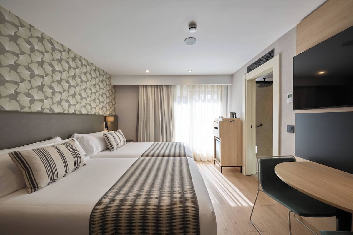 A hotel room with two beds, a desk, a TV, and a shower area.