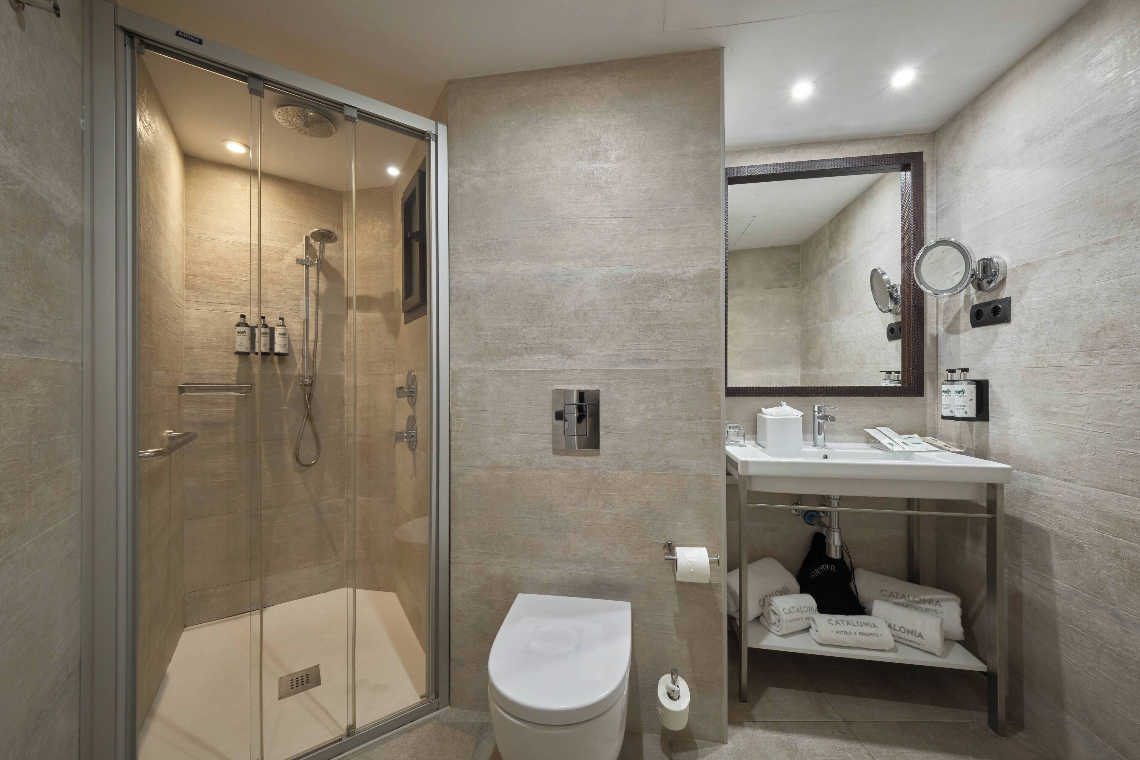 Ein modernes Badezimmer mit einer Glaskabine zur Dusche, einer weißen Toilette, einem Waschbecken mit Spiegel und Pflegeprodukten auf einem Regal.