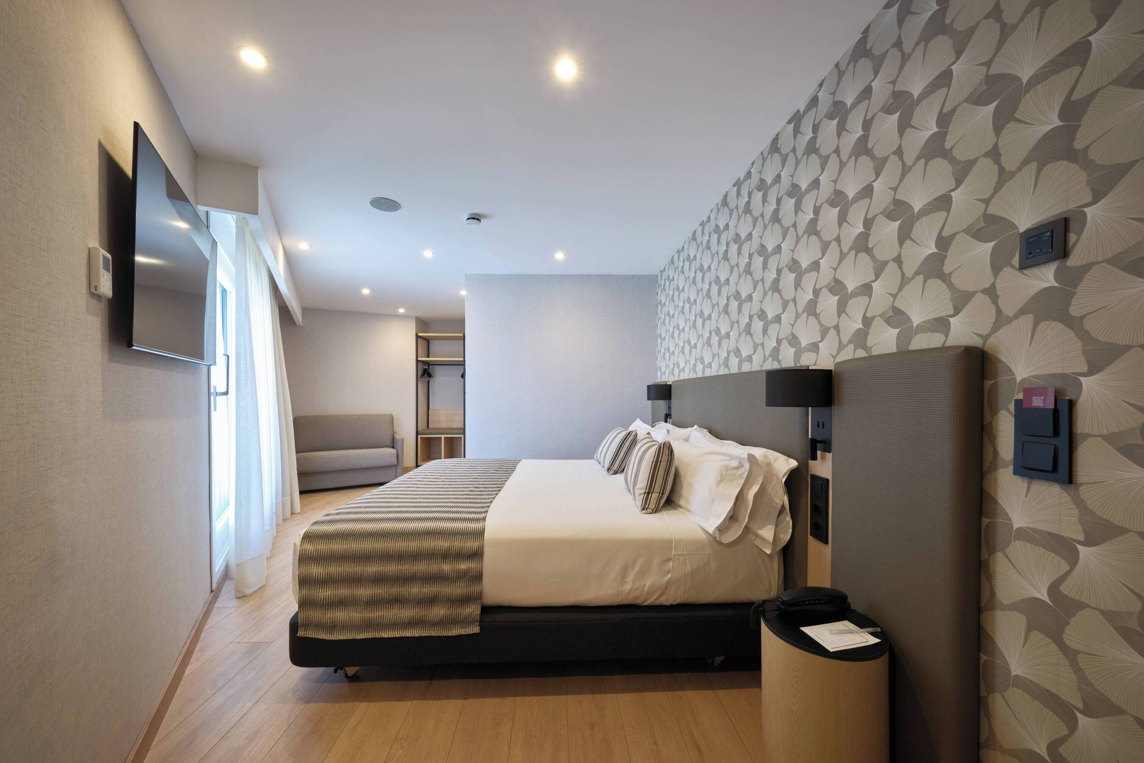 A modern hotel room with a large bed, a flat-screen TV mounted on the wall, a bedside table with a lamp, and a patterned wall.