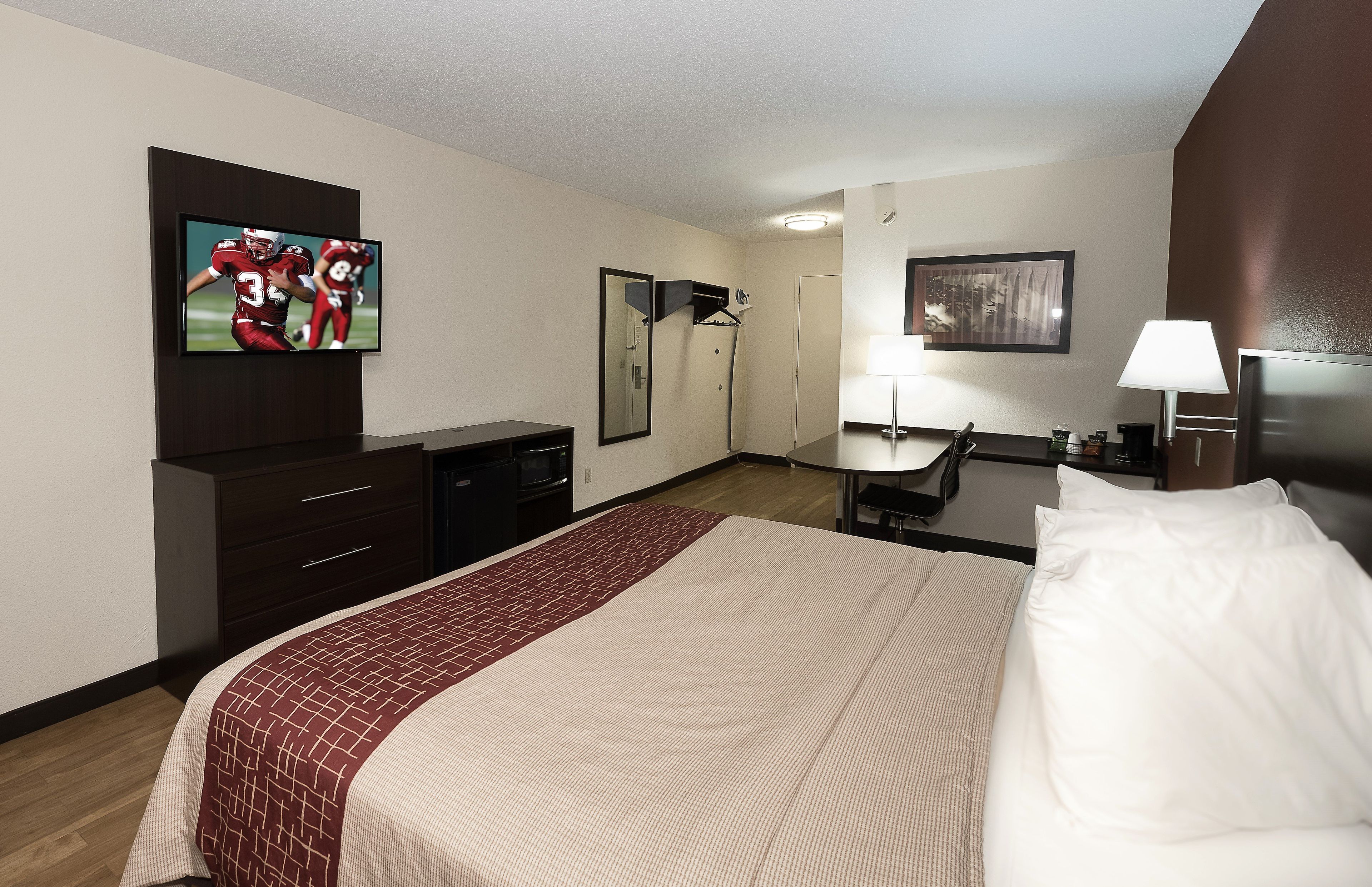 Habitación de hotel con una cama, televisor montado en la pared, escritorio con silla, espejo y lámpara.