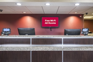 Ein Hotelrezeptionstresen mit einem Flachbildfernseher, der 'Kostenloses WLAN in allen Zimmern' anzeigt, und zwei Computermonitoren.