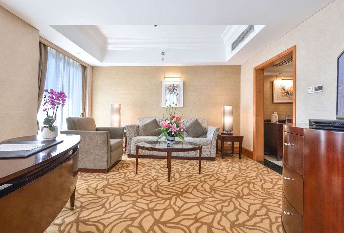 A hotel room with a sofa, armchair, coffee table, and a desk with a lamp.