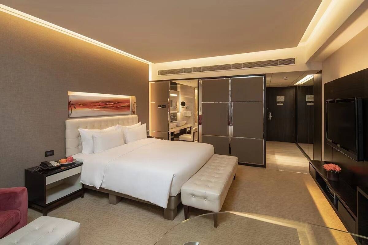 A modern hotel room with a large bed, a desk with a phone, a TV, and a walk-in closet.
