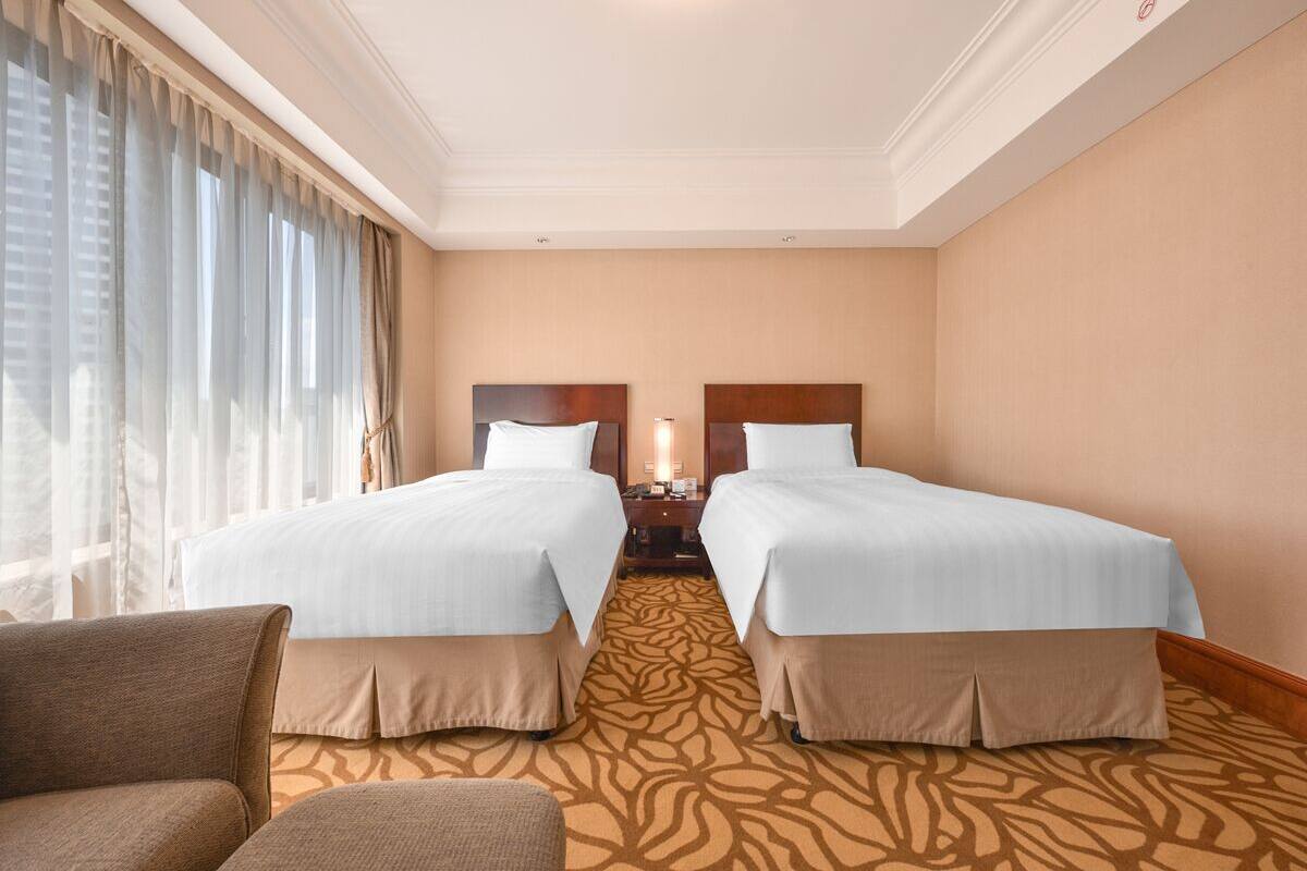A hotel room with two beds, a nightstand, a lamp, and a view of the city through large windows.