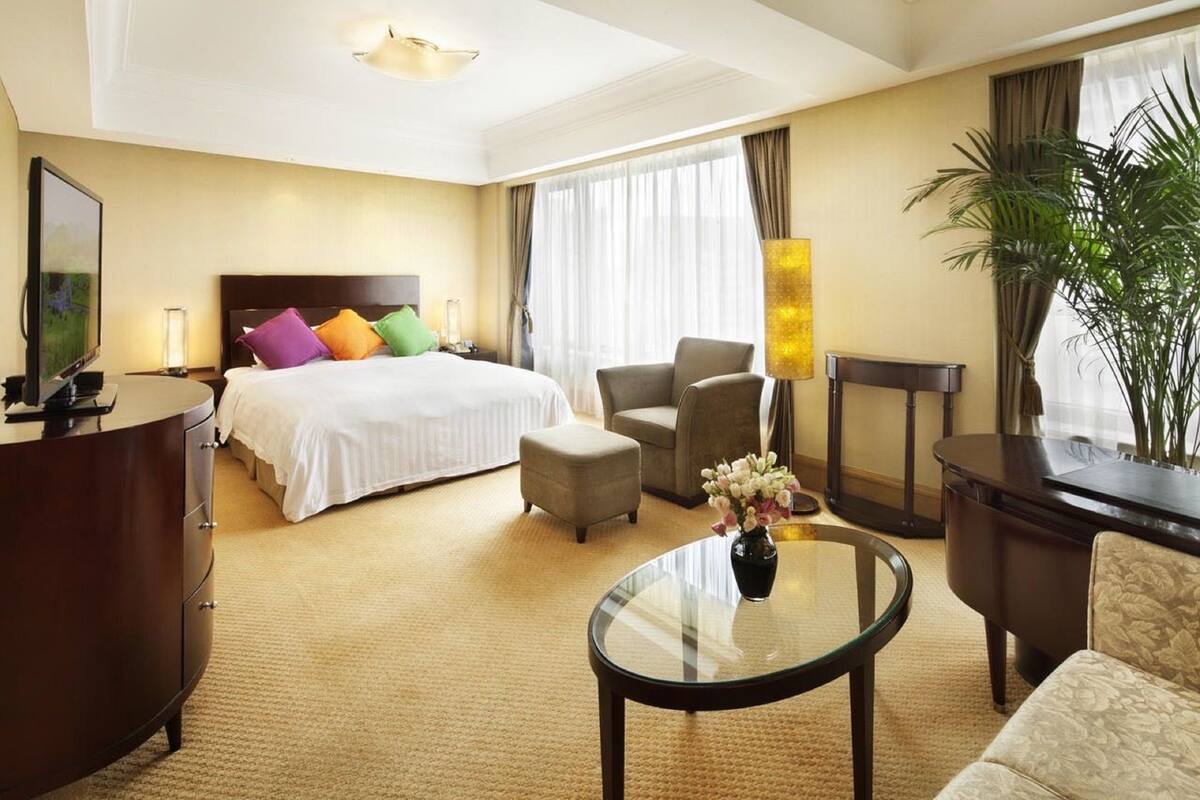 A hotel room with a bed, a desk, a chair, a round table, and a potted plant.