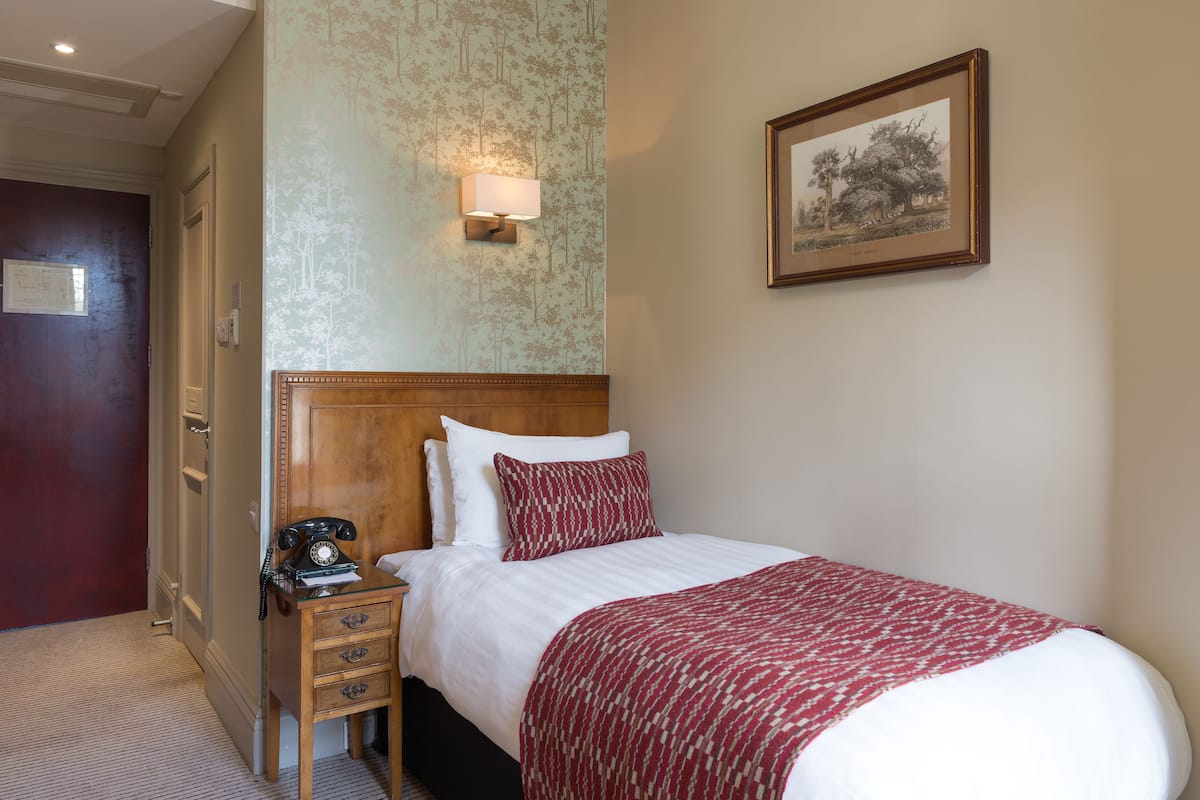 Une chambre d’hôtel avec une tête de lit en bois, un lit simple avec une literie blanche et un plaid à motifs rouges, une table de chevet avec un téléphone à cadran, une lampe fixée au mur et une illustration botanique encadrée.