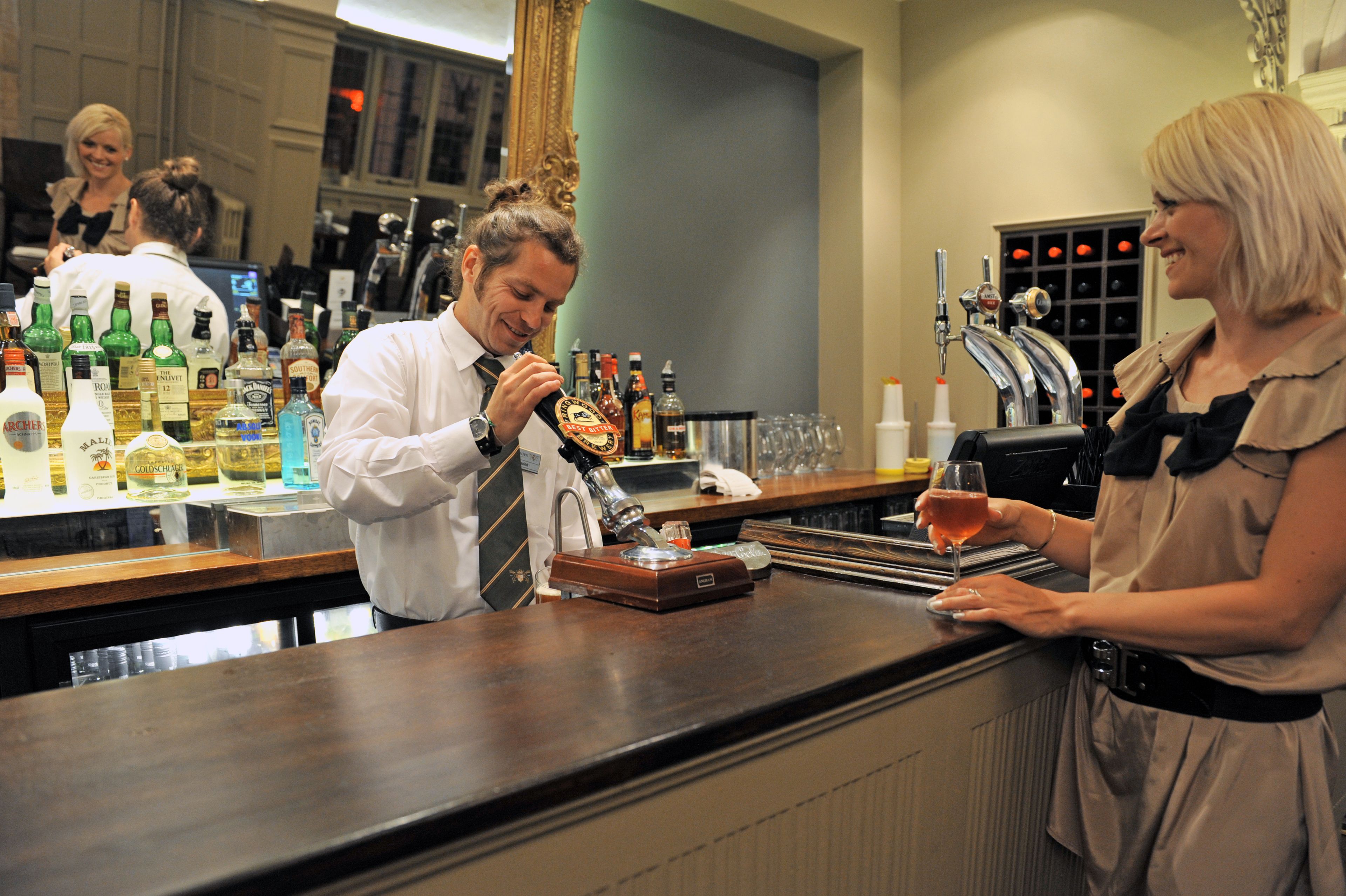 A bar area with a bartender preparing a drink, a wine glass, and various bottles of alcohol on the counter.