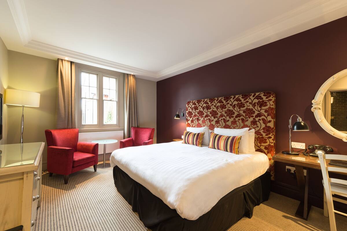 Une chambre d’hôtel avec un grand lit, un bureau avec une lampe, une chaise, une télévision et une fenêtre avec des rideaux.