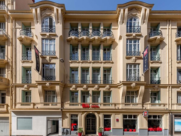 Un hôtel à plusieurs étages, de style architectural classique, doté de balcons avec des garde-corps décoratifs, de grandes fenêtres et d’une entrée imposante surmontée d’une enseigne rouge.