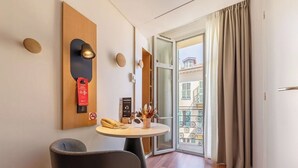 Habitación de hotel con pared de madera decorativa, una mesa redonda con teléfono y un jarrón de flores, una silla y vistas a un balcón con barandilla de hierro forjado.