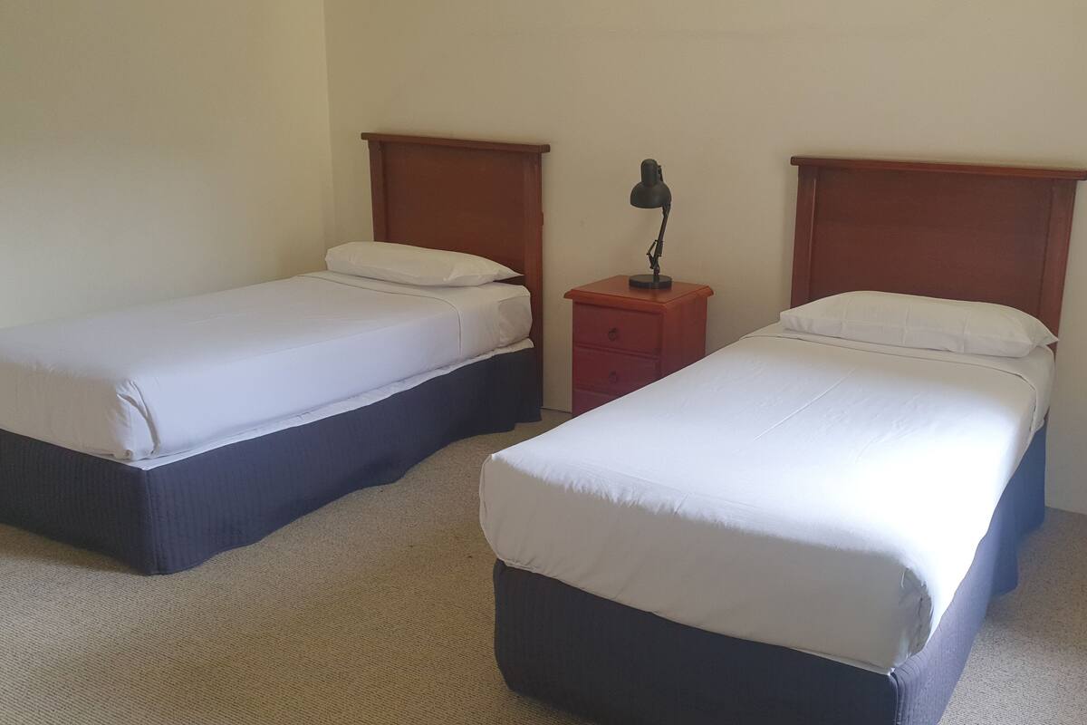Une chambre d’hôtel avec deux lits simples, une table de chevet avec une lampe et un mur sobre.