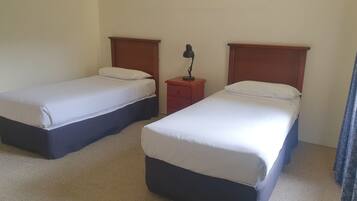 A hotel room with two single beds, a nightstand with a lamp, and a plain wall.