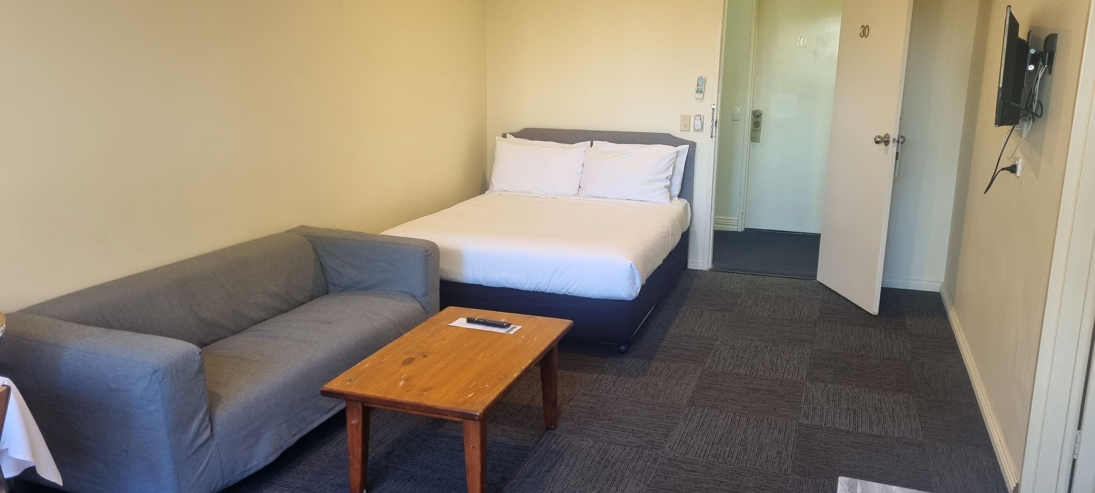 A small, neatly arranged room with a bed, a grey sofa, and a wooden table.