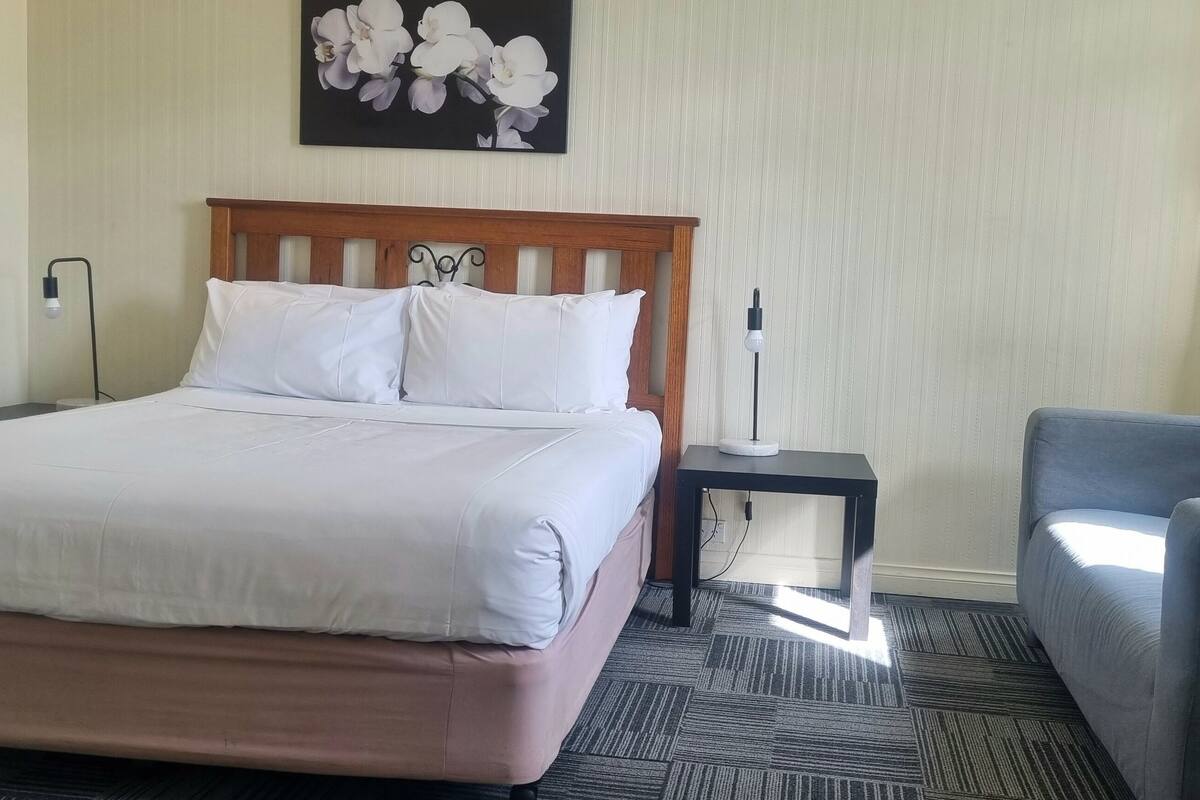 Une chambre d’hôtel comprenant un lit, une table de chevet, une lampe, une chaise et une œuvre d’art accrochée au mur.