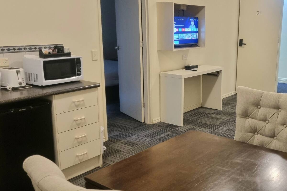 Une chambre d’hôtel compacte comprenant un coin cuisine, un téléviseur à écran plat fixé au mur, une table à manger avec quatre chaises et un petit canapé.