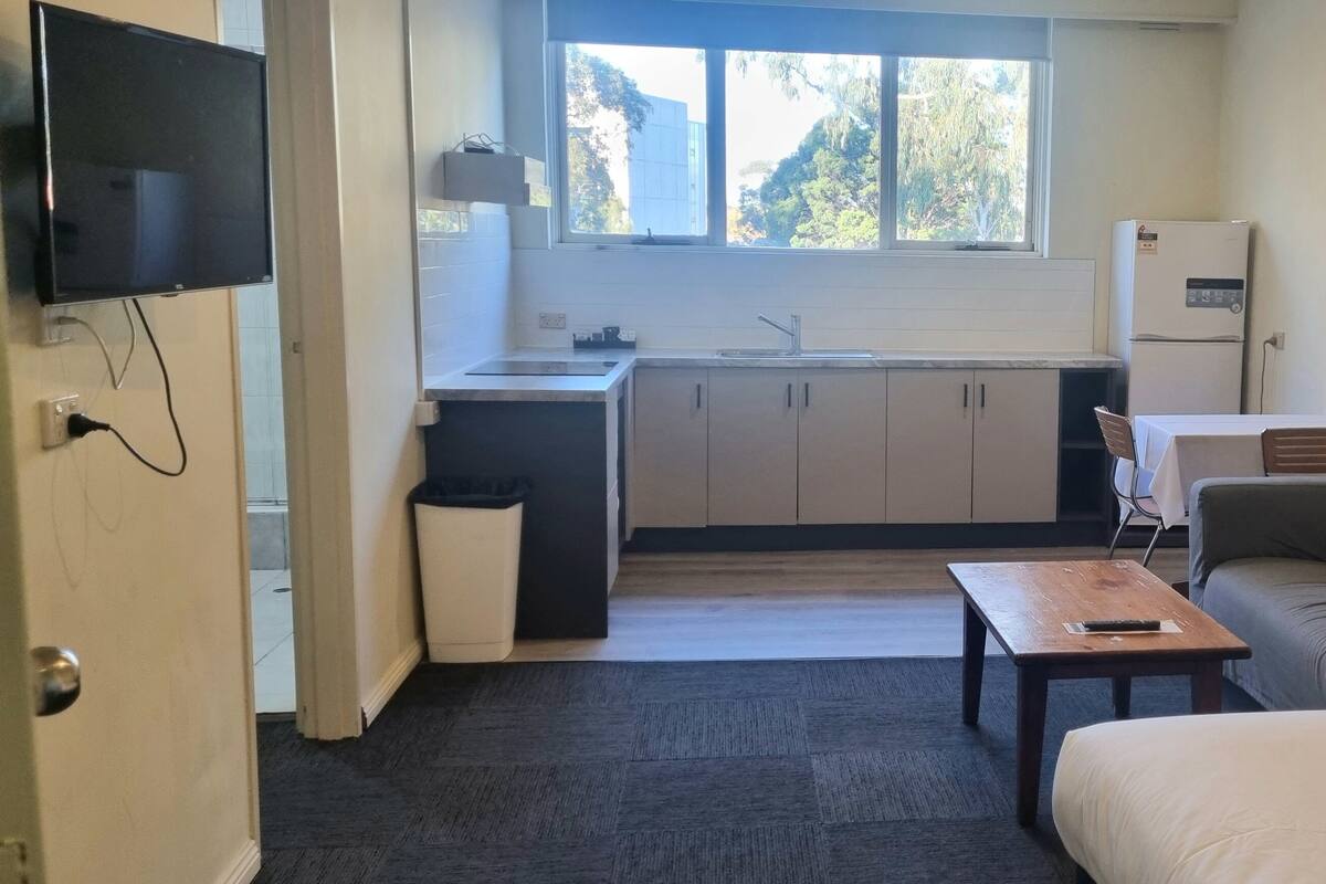 Une pièce compacte comprenant un coin cuisine, un canapé, une petite table et un téléviseur fixé au mur.
