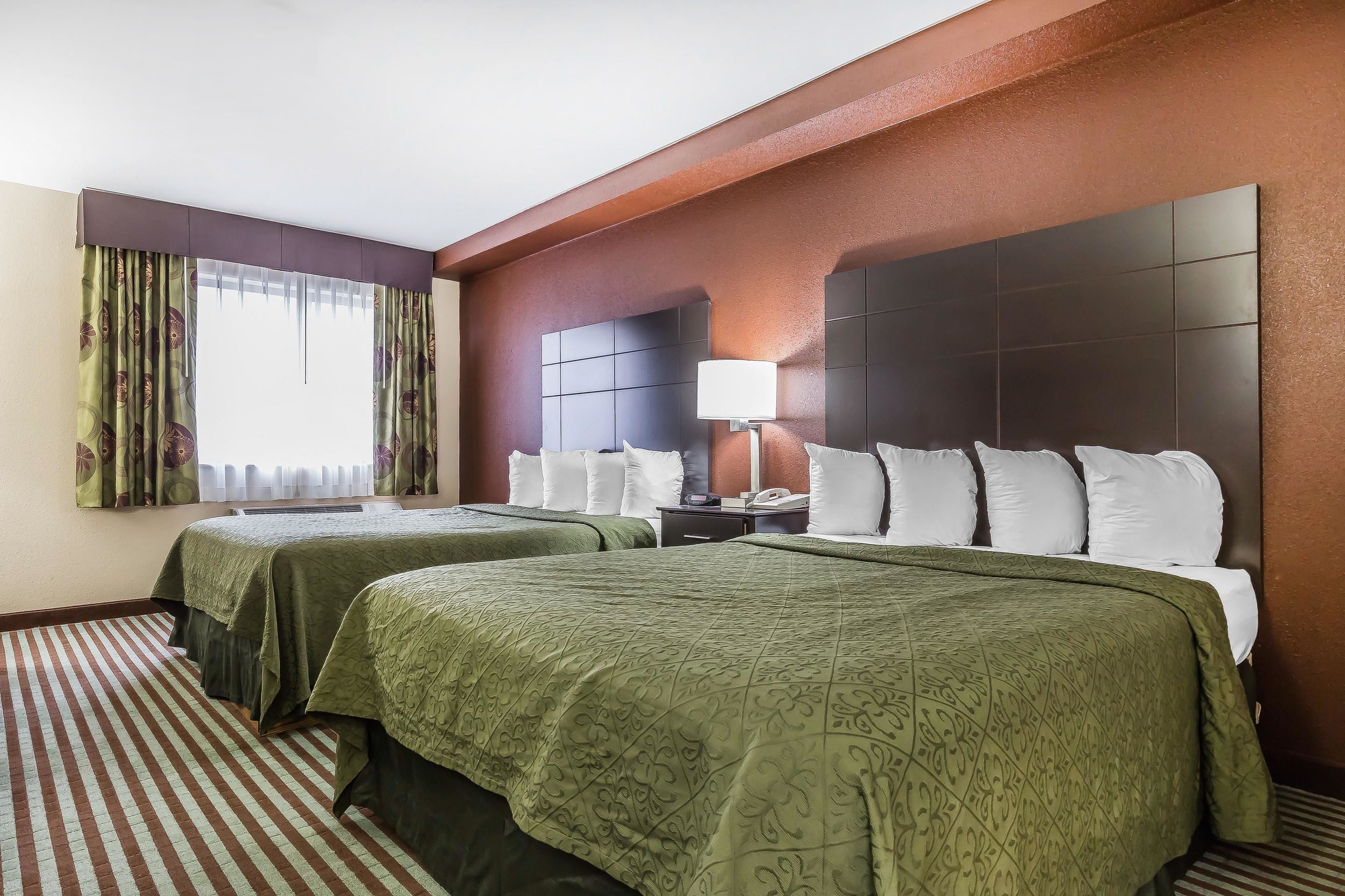 A hotel room with two beds, a large headboard, a bedside table with a lamp, and a window with curtains.