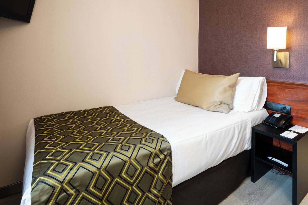 Una habitación de hotel con una cama, una mesita de noche con teléfono y una lámpara de pared.