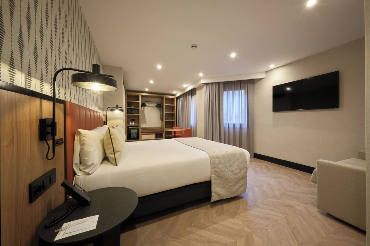 Una habitación de hotel con una cama grande, un escritorio con teléfono, un televisor de pantalla plana montado en la pared y un armario con estantes integrados.