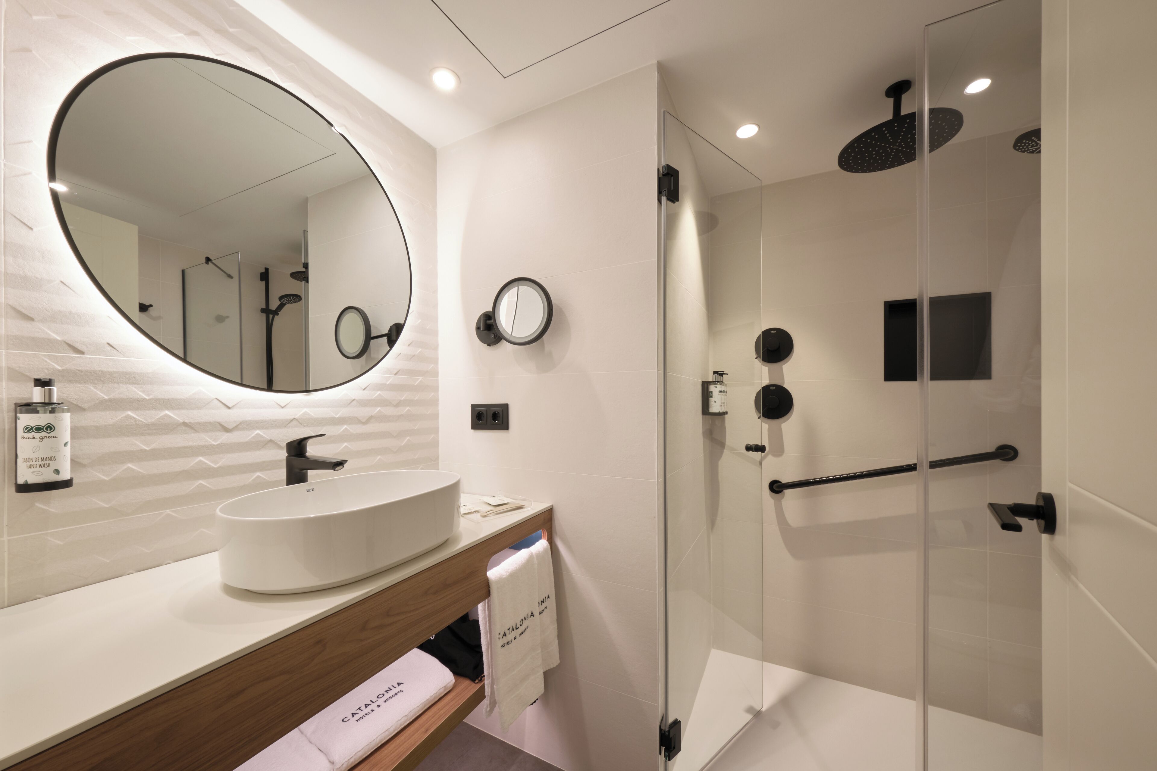A modern bathroom with a large oval mirror, a white sink, and a glass-enclosed shower area.