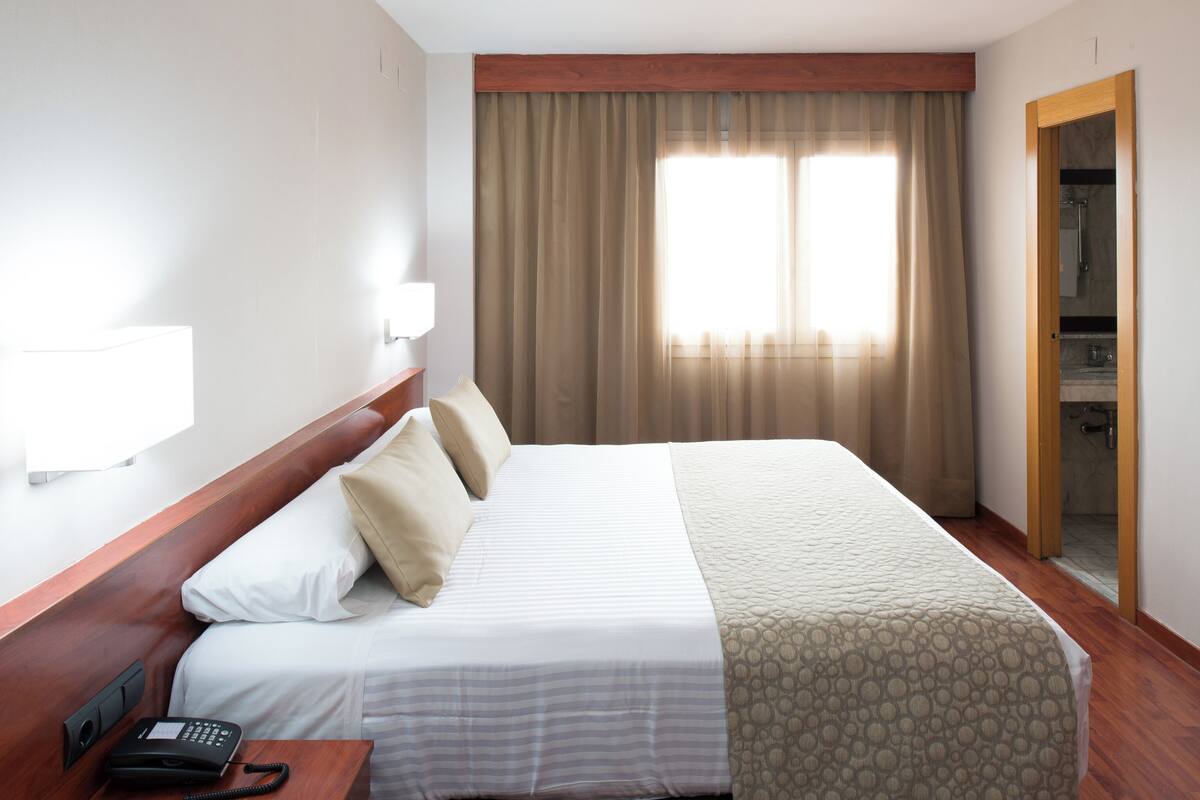 Una habitación de hotel con cama, teléfono, lámpara y ventana con cortinas.
