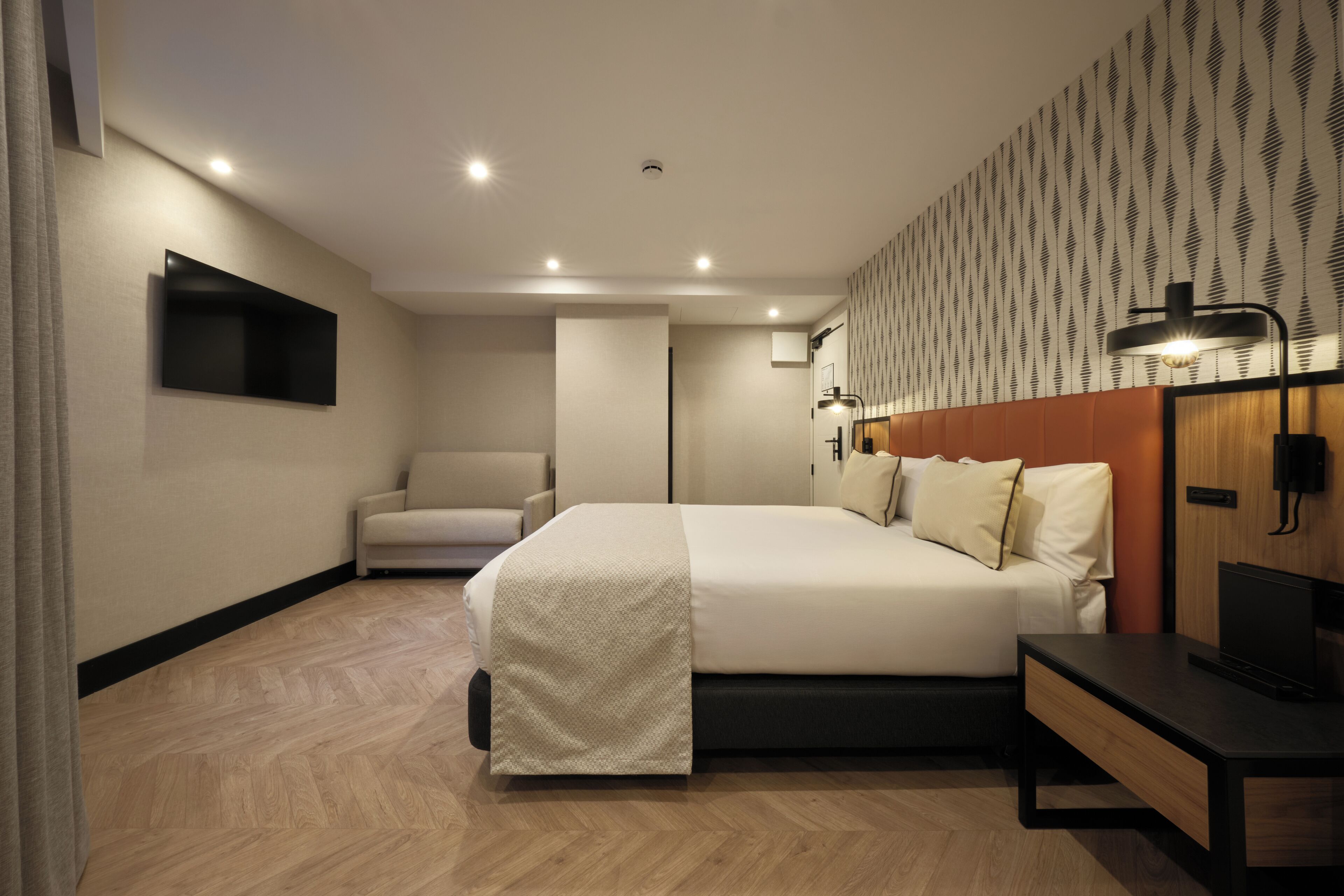 A modern hotel room with a large bed, a desk, a chair, a TV mounted on the wall, and a patterned wallpaper.