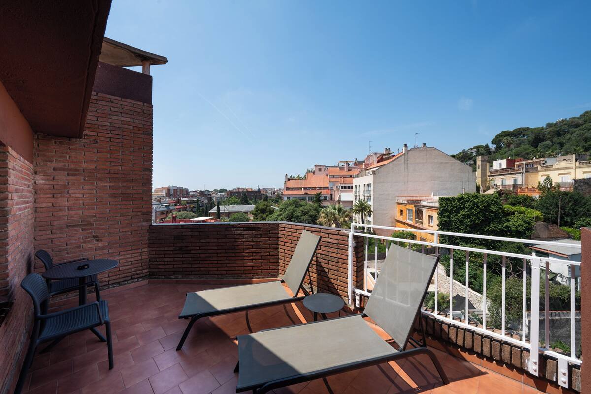 Un balcón con dos sillones de descanso y una mesita, con vistas a un paisaje urbano con edificios y zonas verdes.