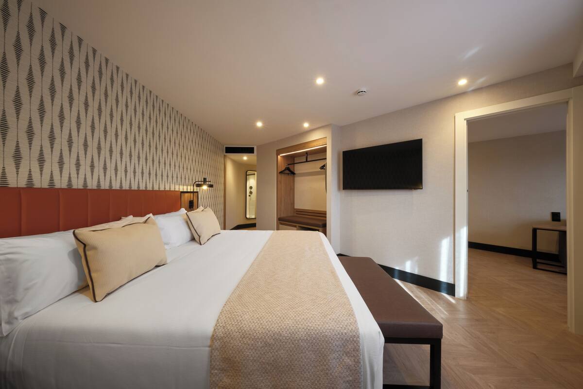 Habitación de hotel con una cama grande, una mesita de noche, un banco, un armario y un televisor montado en la pared.